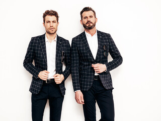 Portrait of two handsome confident stylish hipster lambersexual models. Sexy modern men dressed in black elegant suit. Fashion male posing in studio near white wall