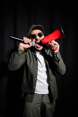 emotional indian hip hop performer in sunglasses and cap screaming in microphone and loudspeaker on black.