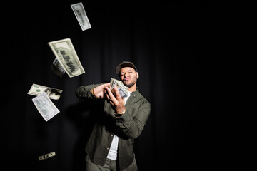 rich indian hip hop performer in cap throwing blurred dollar banknotes on black.