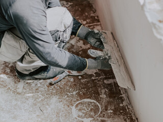 A young caucasian guy is squatting applying fresh putty with a large spatula on the wall