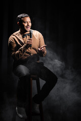 indian comedian sitting and performing stand up comedy into microphone on black with smoke.