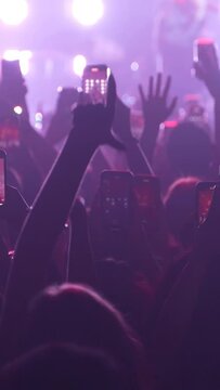 Vertical video of unrecognizable fans dancing at a concert or festival party. Silhouettes of concert crowd in front of bright stage lights