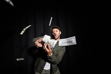 wealthy indian hip hop performer in cap throwing blurred dollar banknotes on black.