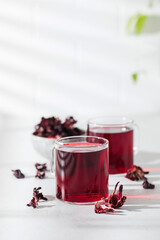 Herbal tea made from hibiscus petals in a transparent cup on a light background and a bowl of dry hibiscus. © Виктория Попова