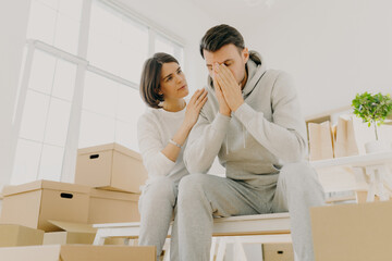 Fototapeta na wymiar Photo of dissatisfied husband and wife forced to sell apartment because of financial problems, woman soothes husband, pose on cardboard boxes, pose in living room. Moving as kind of stress for family