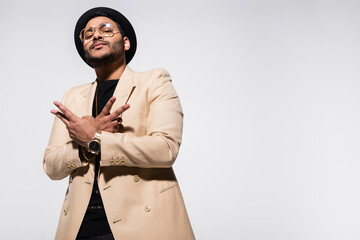 low angle view of indian hip hop performer in fedora hat and eyeglasses gesturing isolated on grey.