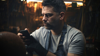A male craftsman in a garage communicating with customer online by smartphone and discussing...
