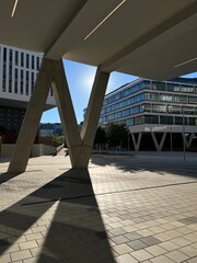 The space under the overhanging corner of a modern office building in the city campus. The sun shines into the underpass and casts a harsh shadow over the massive column onto the V-shaped sidewalk.