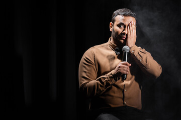 indian comedian covering eye and performing stand up comedy with microphone on black.