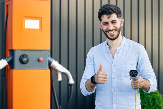 Male hand showing thumbs up holding power cable supply plugged at electric car charging station. Bearded man standing near electric charging station looking at camera and showing thumb up
