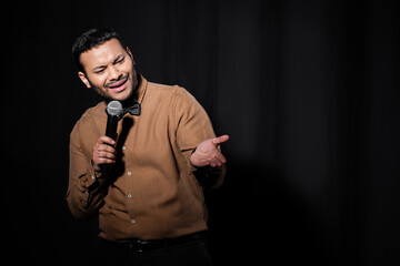 indian comedian in shirt and bow tie holding microphone and talking during performance on black.