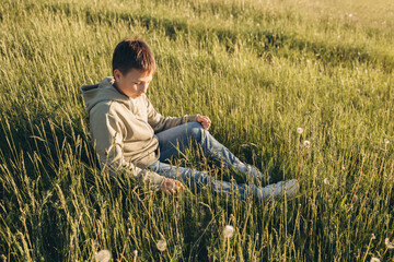 Naklejka na ściany i meble Portrait of happy boy sitting on top of hill in grass field and enjoying beautiful landscape at sunset. Teenager hiker resting in nature. Active lifestyle. Concept of local travel