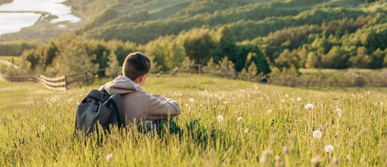 Tourist with backpack sitting on top of hill in grass field and enjoying beautiful landscape view....