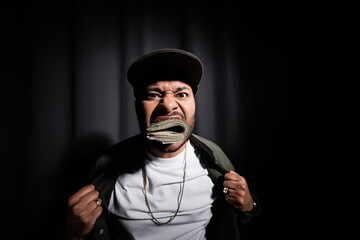 indian hip hop performer in cap biting bundle of dollar banknotes on black.