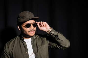 eastern hip hop performer in cap adjusting sunglasses on black.
