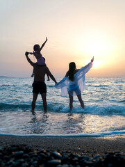 A happy hamily of three members standing in the sea with their hands up and looking at sunset 