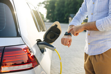 Male hand inserts power connector into EV car and charges batteries, uses smartwatch for activates...