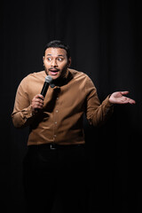 emotional indian comedian in shirt and bow tie holding microphone and gesturing during monologue on black.