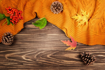 Orange knitted blanket, autumn leaves and cones on a wooden background.
