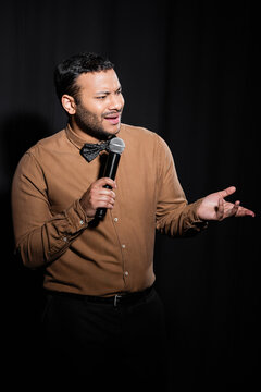 displeased indian stand up comedian in shirt and bow tie holding microphone during monologue on black.