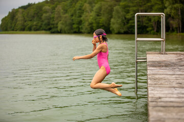 Little sporty girl jumping into water of lake from wooden pier on hot sunny summer day