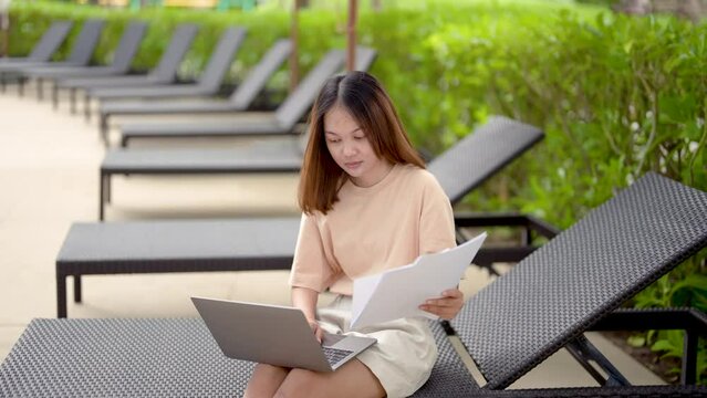 4K, medium shot A woman working at the swimming pool with her laptop computer holding paperwork in her hand. It is imperative for young Asian female tourists to sit and work on vacations at the hotel.