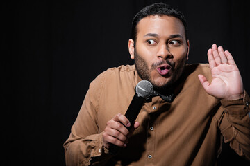 emotional indian stand up comedian showing stop gesture while telling jokes into microphone isolated on black. - obrazy, fototapety, plakaty