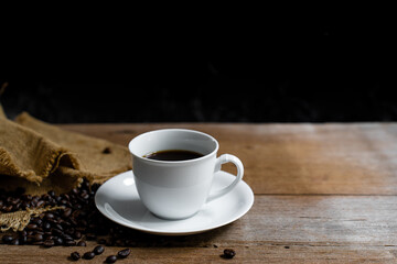 Hot coffee in a white coffee cup and many coffee beans placed around and on a wooden table in a warm, light atmosphere, on dark background, with copy space.