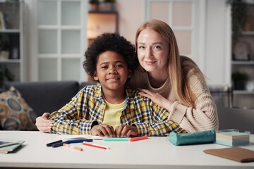 Portrait of young foster mother looking at camera with her adoptive son while they sitting at table...