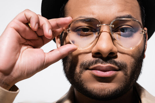 close up of bearded indian hip hop performer in fedora hat adjusting eyeglasses isolated on grey.