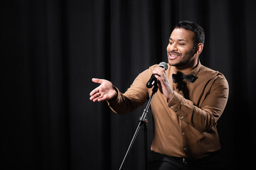 indian comedian performing stand up comedy and telling jokes in microphone while gesturing on black. - obrazy, fototapety, plakaty