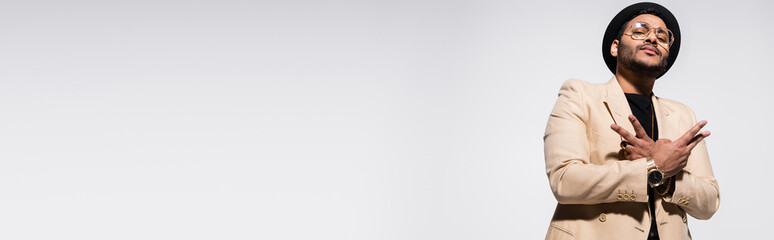 low angle view of indian hip hop performer in fedora hat and eyeglasses gesturing isolated on grey,...