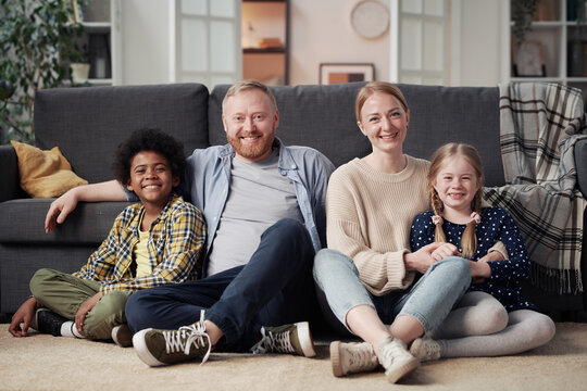 Portrait of happy family with adopted children sitting on floor in living room and smiling at camera