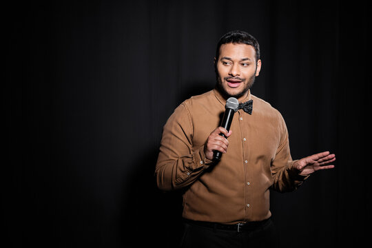 indian stand up comedian with bow tie holding microphone during monologue on black.
