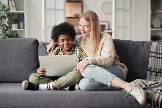 Young mother watching cartoon on laptop with her little adoptive son while they resting on sofa in living room