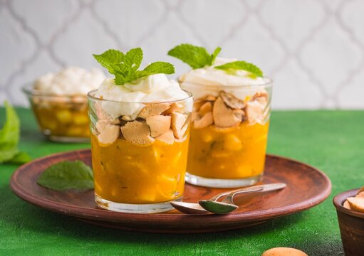 Exotic dessert with mango puree, sliced banana, biscuit cookies and whipped cream in glasses on a green concrete background.