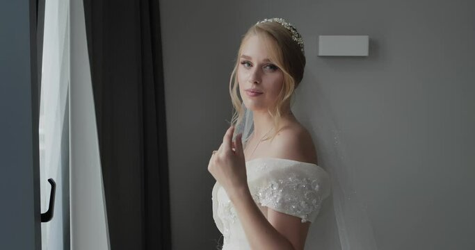 the bride stands by the window in a luxurious wedding dress