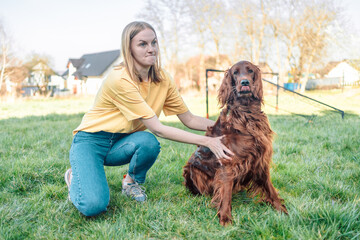 Woman happily smiling at playing with her red Irish setter dog outdoors on fresh green grass in the...