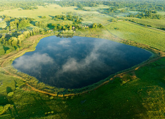 Fototapeta premium Foggy sunrise over lake. Nature at dawn in fog, aerial view. Morning mist haze on lake. Sunny morning scene in misty in rural. Misty landscape on pond.. Sunny foggy hills on sundown. Dawn in hillside.