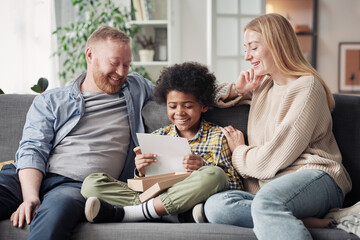Happy child reading a letter about adoption while sitting on sofa with his foster parents