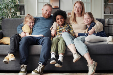 Portrait of big adoptive family with children sitting on sofa, embracing each other and smiling at...