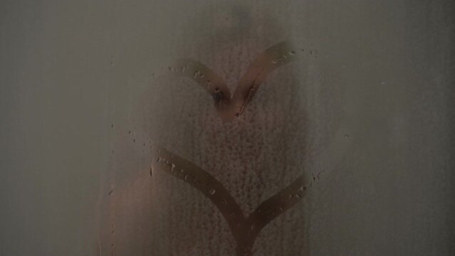 A woman draws a heart on the wet glass in the shower. Drawing of a heart on glass