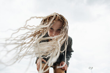 Bending forward freckled teenage girl with dreads against a grey sky.