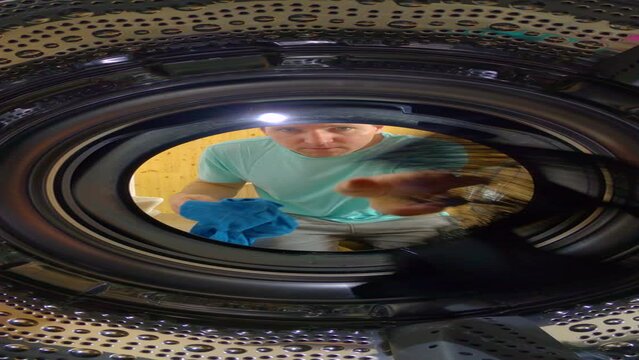 CLOSE UP, VERTICAL, PORTRAIT: Young man loads up the washer with dirty laundry and watches it spin and wash. Curious male doing housework is loading up washing machine and observes it while running.