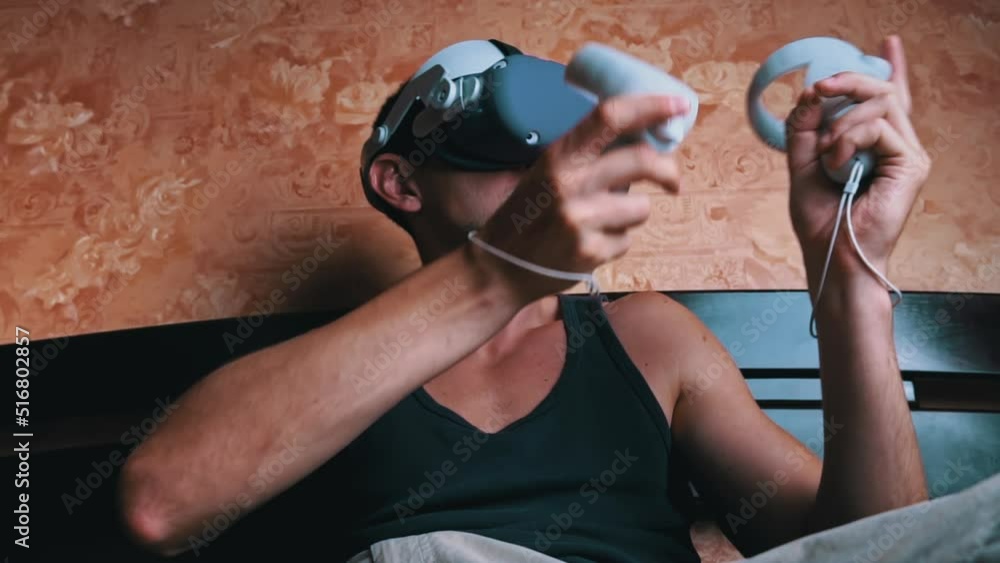 Poster man in a virtual reality helmet controls the controllers. a young man at home sitting on a bed is in
