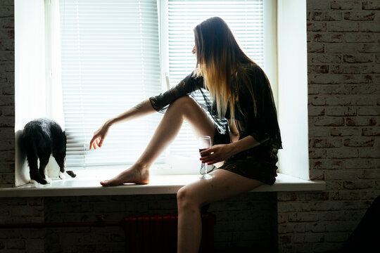 Woman in nightgown sitting on windowsill with glass of wine
