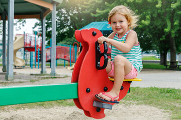 Little child playing at playground in summer, sitting on seesaw swing. Girl playing in park. Happy...