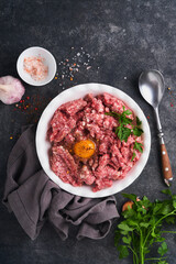 Raw minced meat. Ground meat beef, pork or lamb spices, herbs and eggs on white plate on black old concrete background. Fresh minced meat ready for cooking. Food cooking background. Top view.