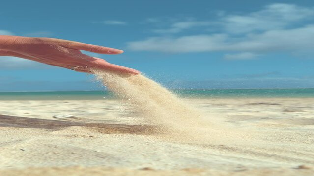 VERTICAL SLOW MOTION, CLOSE UP: Unrecognizable woman lifts up a handful of white sand and lets it fall back down on the calm sandy beach in Cook Islands. Young female on vacation playing with sand.