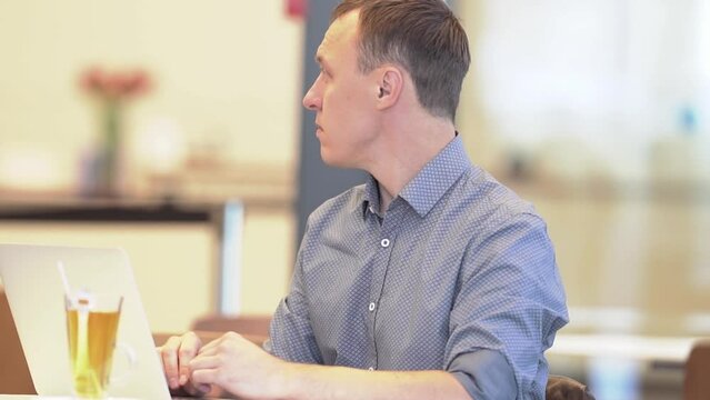 A young man sitting at a table with a laptop in a cafe looks away. A slow-motion average plan of a man. The concept of remote work. High quality FullHD footage